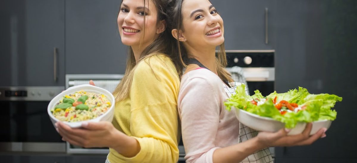 Two smiling women hold vibrant salads in a kitchen setting, showcasing fresh culinary creations.