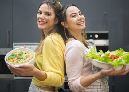 Two smiling women hold vibrant salads in a kitchen setting, showcasing fresh culinary creations.